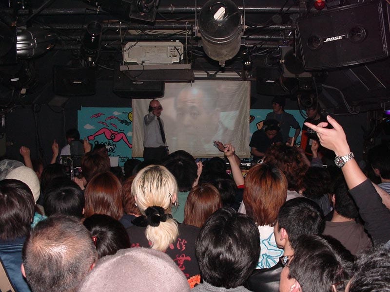 A crowd shot from behind as a DJ performs cosplaying as Muneo Suzuki